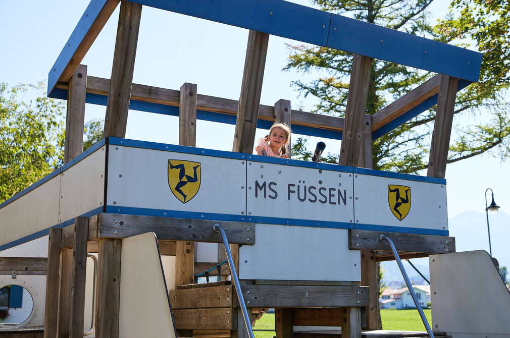 Das Bild fängt ein junges Mädchen ein, das hinter einer hölzernen Konstruktion hervorlugt. Es lacht und scheint sich zu amüsieren. Die hölzerne Konstruktion ist mit einem blauen und weißen Schild verziert, das die Aufschrift "MS FUSSEN" trägt. Das Schild ist an einem hölzernen Rahmen montiert, der durch hölzerne Balken gestützt wird. Der Hintergrund des Bildes ist von Bäumen gefüllt, was darauf hindeutet, dass sich der Ort in einem Park oder einer ähnlichen Außenanlage befinden könnte. Die Position des Mädchens hinter der hölzernen Konstruktion und sein Blick zur Kamera erzeugen ein Gefühl von Neugier und Spielfreude. Die gesamte Szene vermittelt ein Gefühl von Spaß und Abenteuer.