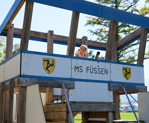 Das Bild fängt ein junges Mädchen ein, das hinter einer hölzernen Konstruktion hervorlugt. Es lacht und scheint sich zu amüsieren. Die hölzerne Konstruktion ist mit einem blauen und weißen Schild verziert, das die Aufschrift "MS FUSSEN" trägt. Das Schild ist an einem hölzernen Rahmen montiert, der durch hölzerne Balken gestützt wird. Der Hintergrund des Bildes ist von Bäumen gefüllt, was darauf hindeutet, dass sich der Ort in einem Park oder einer ähnlichen Außenanlage befinden könnte. Die Position des Mädchens hinter der hölzernen Konstruktion und sein Blick zur Kamera erzeugen ein Gefühl von Neugier und Spielfreude. Die gesamte Szene vermittelt ein Gefühl von Spaß und Abenteuer.