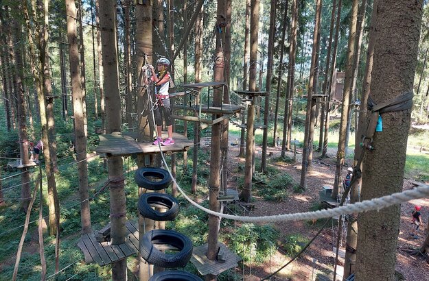  Das Bild fängt eine lebhafte Szene auf einem Spielplatz ein. Der Spielplatz befindet sich inmitten eines Waldes, wobei Bäume und andere Vegeation im Hintergrund in die Höhe ragen. Der Spielplatz ist mit verschiedenen Strukturen ausgestattet, darunter eine Seilbrücke, eine Holzplattform und ein Baumhaus.  In der Vordergrund sieht man ein Kind, welches sich eine Seiltreppe hinaufklettert, wobei ihre Hände sich fest um die Seile schmiegen. Das Abenteuerlust des Kindes ist offensichtlich, während sie die Spielgeräte erkunden.  Im Hintergrund kann man sehen, wie andere Kinder den Spielplatz genießen. Sie sind über den Spielplatz verteilt, einige auf der Seilbrücke, andere auf der Holzplattform und einige im Baumhaus. Ihre fröhliche Aktivität trägt zur lebhaften Atmosphäre des Spielplatzes bei.  Der Spielplatz ist nicht nur für Spielen da, sondern auch ein Ort für Lernen und Erkundung. Es ist der Ort, an dem Kinder ihre physischen Fähigkeiten, soziale Fertigkeiten und Kreativität entwickeln können. Der Spielplatz, mit seinen verschiedenen Strukturen und der umgebenden Vegetation, bietet eine sichere und anregende Umgebung für Kinder zum Wachsen und Lernen.
