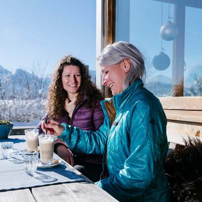 Das Bild zeigt zwei Frauen, die auf einer Terrasse vor einem Haus an einem Holztisch sitzen. Sie heben beide ein Glas Wasser und je ein Glas Latte Macchiato vor sich stehe. Sie tragen Winterkleidung. Eine Frau rührt mit einem Löffel in ihrem Latte Macchiato. Im Hintergrund sieht man die schneebedeckte Umgebung und die verschneiten Berge. Der Himmel ist blau. Die Sonne scheint.  