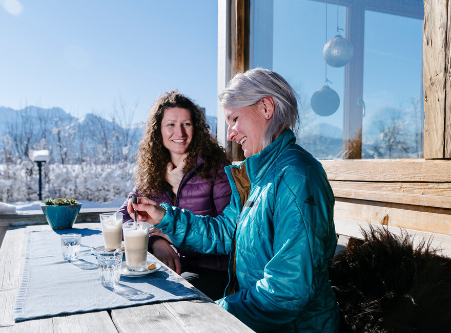 Das Bild zeigt zwei Frauen, die auf einer Terrasse vor einem Haus an einem Holztisch sitzen. Sie heben beide ein Glas Wasser und je ein Glas Latte Macchiato vor sich stehe. Sie tragen Winterkleidung. Eine Frau rührt mit einem Löffel in ihrem Latte Macchiato. Im Hintergrund sieht man die schneebedeckte Umgebung und die verschneiten Berge. Der Himmel ist blau. Die Sonne scheint.  