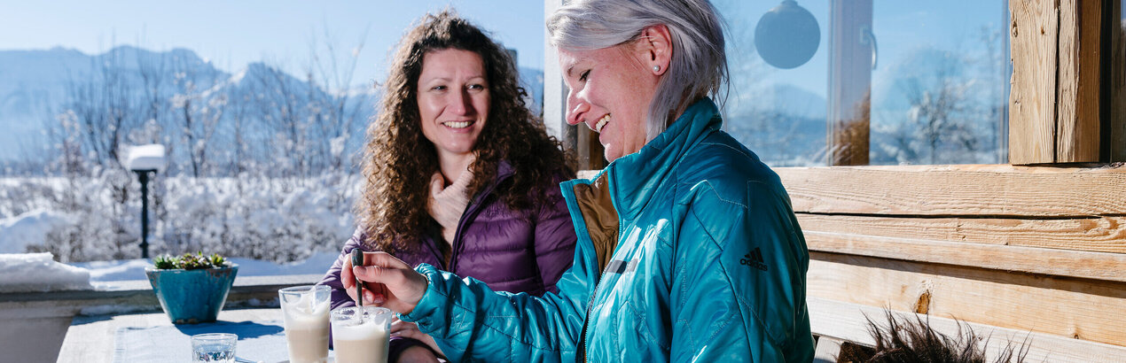 Das Bild zeigt zwei Frauen, die auf einer Terrasse vor einem Haus an einem Holztisch sitzen. Sie heben beide ein Glas Wasser und je ein Glas Latte Macchiato vor sich stehe. Sie tragen Winterkleidung. Eine Frau rührt mit einem Löffel in ihrem Latte Macchiato. Im Hintergrund sieht man die schneebedeckte Umgebung und die verschneiten Berge. Der Himmel ist blau. Die Sonne scheint.  