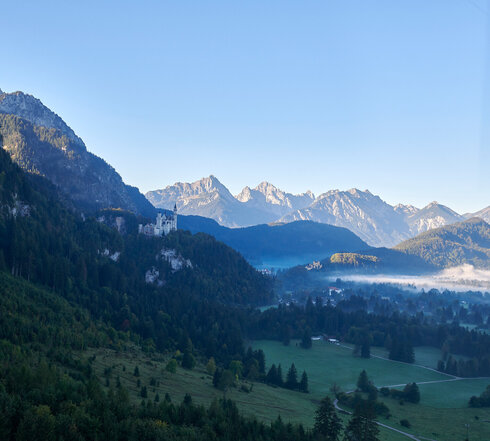 Das Bild zeigt das Märchenschloss Neuschwanstein und das Schloss Hohenschwangau, die im Schatten des Tegelbergs liegen. Auf den grünen Wiesen unterhalb der Schlösser liegen noch Reste des Morgennebels, während die Berggipfel der Allgäuer Alpen im Hintergrund bereits hell von der aufgehenden Sonne angestrahlt werden. Der Himmel ist klar und wolkenlos.