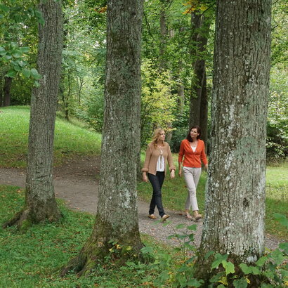 Das Bild zeigt zwei Frauen, die auf einen Feldweg laufen. Ringsum wachsen hohe Bäume. Der Boden ist mit Gras bewachsen. Die Sonne scheint. 