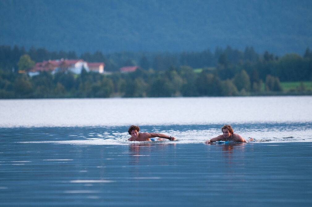 Das Bild zeigt eine friedliche Szene von zwei Männern, die bäuchlings je auf einem Surfbrett liegen und so durch den Hopfensee schwimmen. Der tiefblaue See liegt eingebettet in einen Hintergrund aus grünen Bäumen und Hügeln. Die Männer, beide mit nacktem Oberkörper, befinden sich mitten im Schwimmvergnügen, ihre Körper sind teilweise im Wasser. Ein Mann ist etwas weiter voraus als der andere, beide schwimmen in dieselbe Richtung, als ob sie einem gemeinsamen Ziel entgegenstreben. Die Stille der Szene wird durch die Abwesenheit anderer Personen oder Gegenstände in der Umgebung noch verstärkt. Die Gesamtkomposition des Bildes suggeriert einen friedvollen und entspannten Tag in der Natur.