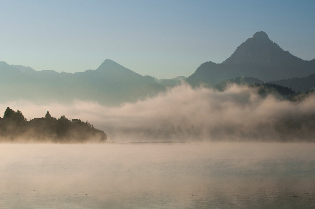 Das Bild zeigt in Nebel gehüllte Berge, die sich majestätisch im Hintergrund erheben.  Der zartweiße Nebel verleiht der Szene einen Hauch von mystischer Stimmung. Im Vordergrund spiegelt der ruhige Weißensee die majestätischen Berge. Über den tiefblauen See ziehen die feinen Nebelschwaden hinweg. Der Himmel darüber ist hell und blau. 