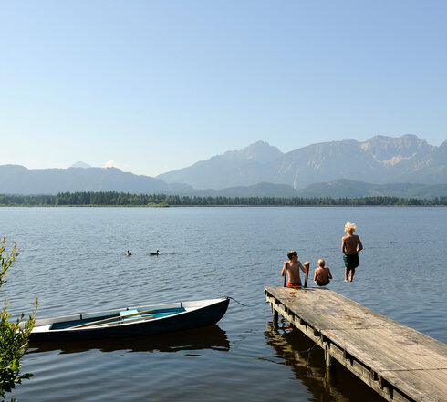 Die Aufnahme erfasst eine idyllische Szenerie am Hopfensee. Im Vordergrund erstreckt sich ein hölzerner Steg ins Wasser, an dem sich drei Kinder tummeln. Sie springen vom Steg ins Wasser oder klettern gerade die Leiter aus dem Wasser zurück auf den See. Der Steg ist auf der rechten Seite des Bildes zu sehen. Im Hintergrund erheben sich majestätisch Berge, deren Spitzen den klaren blauen Himmel berühren. Die Berge bilden eine atemberaubende Kulisse für den ruhigen See. Links vom Bild liegt ein kleines blaues Boot am Steg angedockt. 