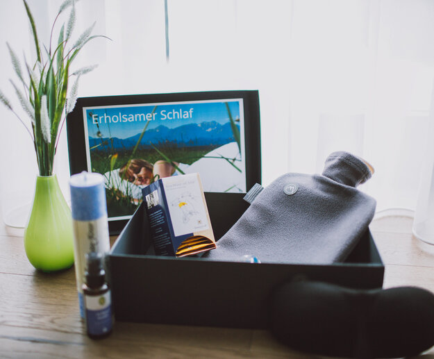 Das Bild zeigt unterschiedliche Gegenstände, die auf dem Holzboden vor einem lichtdurchfluteten Stohr stehen. Von links nach rechts: Eine bauchige grüne Vase mit Gräsern darin. Ein Bilderrahmen enthält das Bild einer Frau, die mit Kopfkissen und Decke in einer Wiese schläft, eine Packung Teebeutel, eine Wärmflasche, eine hohe Kerze, eine kleine braune Sprayflasche und eine schwarze Augenmaske. 