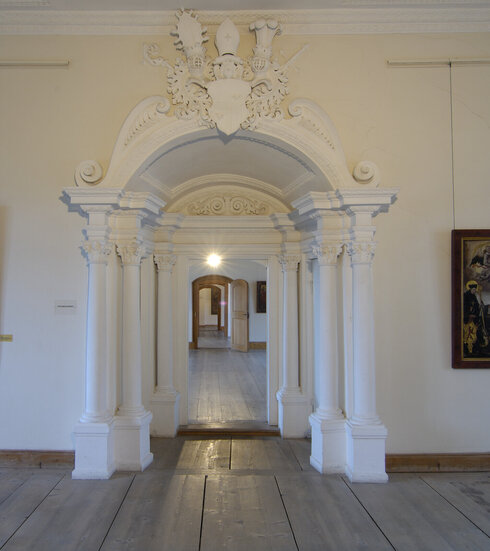 Das Bild zeigt ein mit weißem Stuck reich verziertes Portal. Das von sechs weißen Säulen flankiert ist. Das Portal führt in einen weiteren Raum. Der Boden ist mit einem historischen schlichten Holzboden ausgelegt. Die Wände sind weiß. Auf den Wänden im Vordergrund hängen historische Gemälde.  