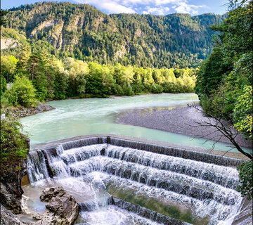 Das Bild zeigt einen atemberaubenden Blick auf einen Wasserfall – den Lechfall bei Füssen, der sich über eine felsige Klippe hinunterstürzt. Der Wasserfall, mit seinem weißen Schaum, ist der Hauptakzent im Bild. Er ist von üppig grünen Bäumen und Büschen umgeben, die der Szene eine Prise der Naturpracht verleihen. Der Wasserfall liegt in einem Tal, durch das der Fluss Lech fließt. Der klare blaue Himmel über ihm ist von fluffigen, weißen Wolken durchzogen. Die Perspektive des Bildes ist aus einer hohen Position, was eine Panoramaansicht des Wasserfalls und seiner Umgebung bietet.