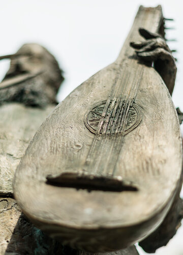 Ein Detail einer Bronzestatue ist zu sehen. Man sieht einen bärtigen Herrn, der eine Laute hält. Die Laute steht im Fokus. Der Rest des Bildes ist unscharf. 