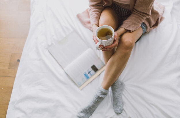 Das Bild zeigt eine Frau, die mit gekreuzten Beinen auf einem weiß bezogenen Bett sitzt. Sie hält eine Teetasse in der Hand. Neben ihr liegt ein Buch im Bett. Mann kann ihren Kopf nicht sehen.