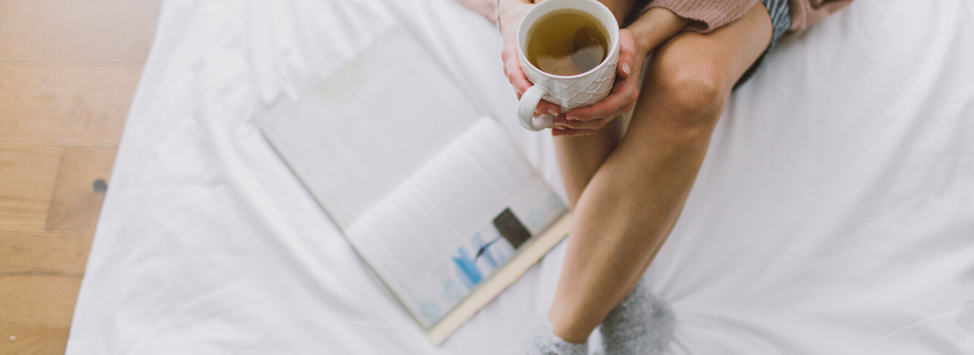 Das Bild zeigt eine Frau, die mit gekreuzten Beinen auf einem weiß bezogenen Bett sitzt. Sie hält eine Teetasse in der Hand. Neben ihr liegt ein Buch im Bett. Mann kann ihren Kopf nicht sehen.