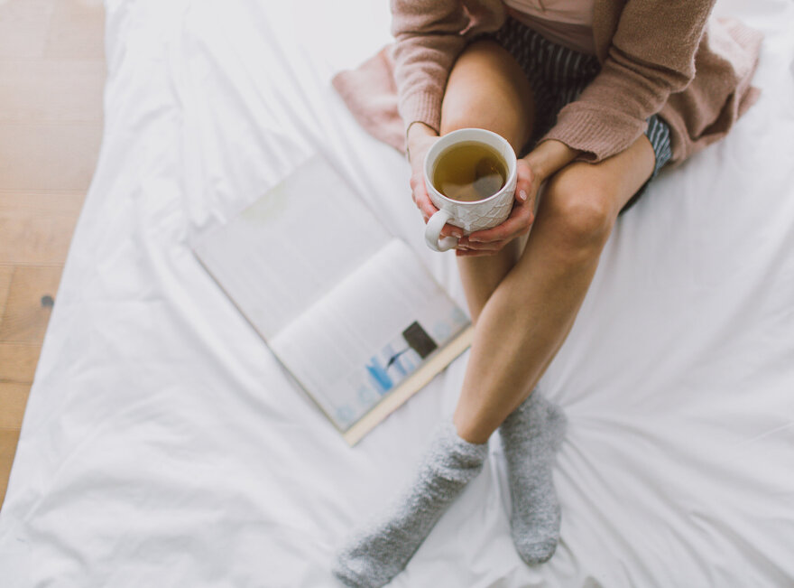 Das Bild zeigt eine Frau, die mit gekreuzten Beinen auf einem weiß bezogenen Bett sitzt. Sie hält eine Teetasse in der Hand. Neben ihr liegt ein Buch im Bett. Mann kann ihren Kopf nicht sehen.