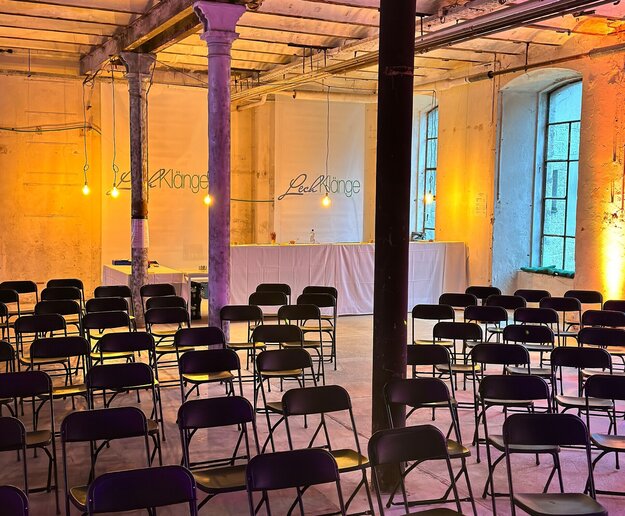Das Bild zeigt einen Raum, der von drei Säulen unterbrochen wird. An der Hinterwand des Raumes hängen lange Banner auf denen „Lechklänge“ zu lesen ist. Davor ist ein Tresen zu erkennen. Der rosabeleuchtete Raum ist fast vollständigen mit Stühlen in Kinobestuhlung gefüllt.