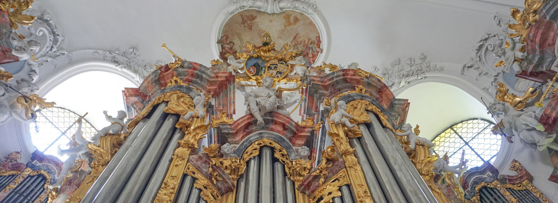 Das Bild zeigt die Pfeifen einer Kirchenorgel, die mit buntem Stuck ringsum reich verziert sind. Das Foto ist von unten aufgenommen.  