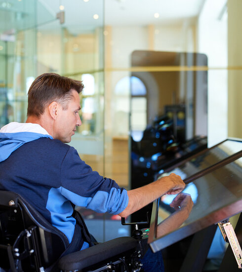 Das Bild zeigt einen Mann in einem Rollstuhl, der sich vollständig in das Spielen eines Videospiels vertieft. Er sitzt bequem in seinem Rollstuhl, welcher sich direkt vor einem großen Bildschirm befindet. Der Mann hält einen Spielcontroller in der Hand, seine Finger fähig, die Knöpfe geschickt zu bedienen. Der Bildschirm vor ihm zeigt ein lebhaftes Spiel voller Farben und Grafiken. Der Mann ist vollständig auf das Spiel konzentriert, seine Aufmerksamkeit unerschütterlich. Die Kulisse scheint ein Raum mit einer Glaswand zu sein, welche genügend natürliches Licht durchlässt. Die Atmosphäre ist eine von Konzentration und Vergnügen.