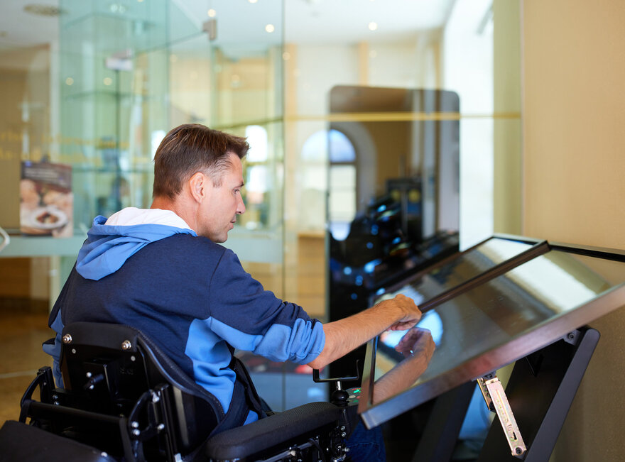 Das Bild zeigt einen Mann in einem Rollstuhl, der sich vollständig in das Spielen eines Videospiels vertieft. Er sitzt bequem in seinem Rollstuhl, welcher sich direkt vor einem großen Bildschirm befindet. Der Mann hält einen Spielcontroller in der Hand, seine Finger fähig, die Knöpfe geschickt zu bedienen. Der Bildschirm vor ihm zeigt ein lebhaftes Spiel voller Farben und Grafiken. Der Mann ist vollständig auf das Spiel konzentriert, seine Aufmerksamkeit unerschütterlich. Die Kulisse scheint ein Raum mit einer Glaswand zu sein, welche genügend natürliches Licht durchlässt. Die Atmosphäre ist eine von Konzentration und Vergnügen.