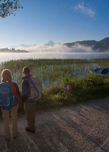 Das Bild zeigt drei Personen in Wanderkleidung und mit Rucksäcken, die am Ufer eine Sees stehen. Sie blicken auf das Wasser und haben dem Betrachter den Rücken zugedreht. Über dem Wasser wabern noch Nebelschwaden. Die Sonne scheint der Himmel ist blau. Im Hintergrund sieht man hohe Berge und bewaldete Hügel.    