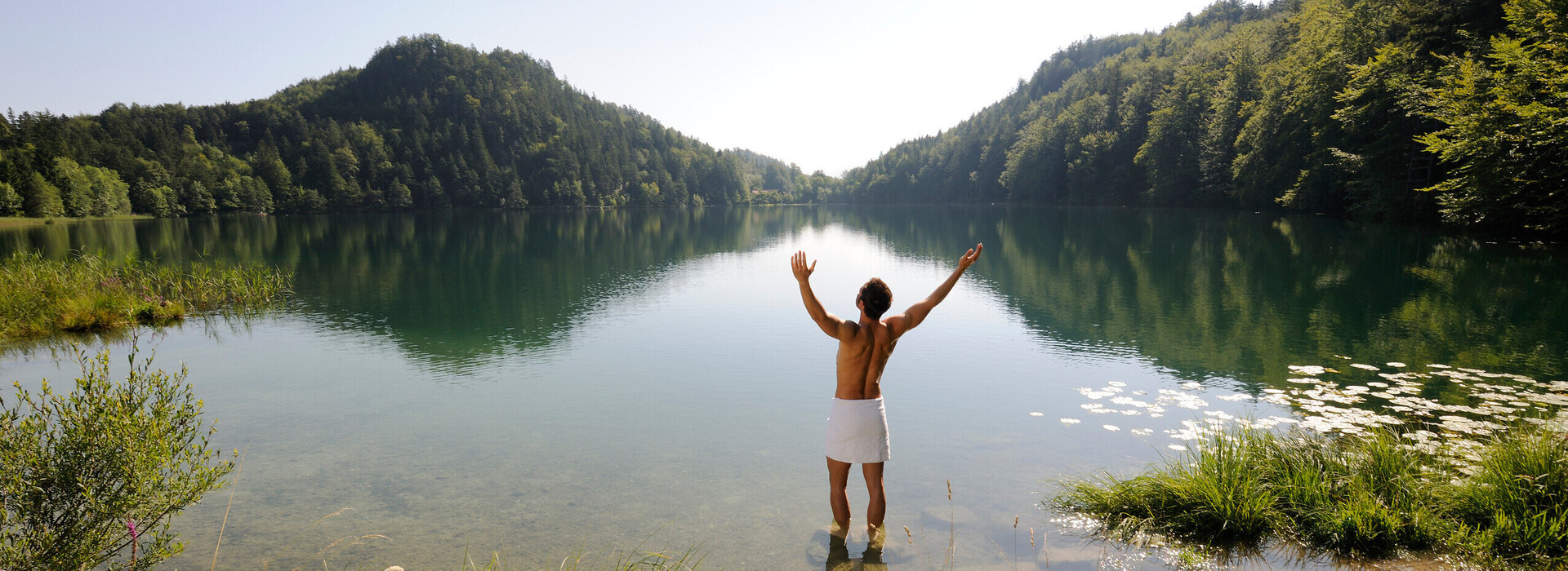 Das Bild zeigt einen Mann, der sich nur ein weißen Handtuch um die Hüften gebunden hat. Er hat dem Betrachter den Rücken zugedreht, steht im knöcheltiefen Wasser eines Sees und streckt die Arme nach oben. Der See ist umringt von grünen Wäldern. Der Himmel ist blau. Die Sonne scheint.        