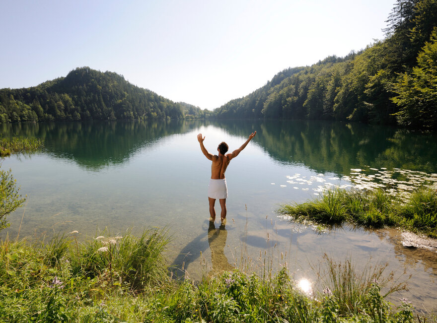 Das Bild zeigt einen Mann, der sich nur ein weißen Handtuch um die Hüften gebunden hat. Er hat dem Betrachter den Rücken zugedreht, steht im knöcheltiefen Wasser eines Sees und streckt die Arme nach oben. Der See ist umringt von grünen Wäldern. Der Himmel ist blau. Die Sonne scheint.        
