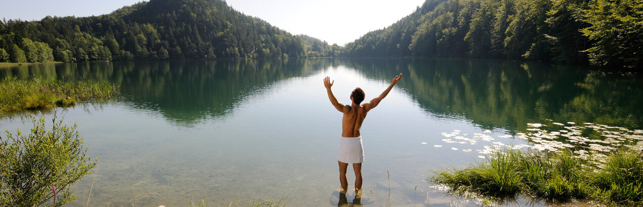 Das Bild zeigt einen Mann, der sich nur ein weißen Handtuch um die Hüften gebunden hat. Er hat dem Betrachter den Rücken zugedreht, steht im knöcheltiefen Wasser eines Sees und streckt die Arme nach oben. Der See ist umringt von grünen Wäldern. Der Himmel ist blau. Die Sonne scheint.        