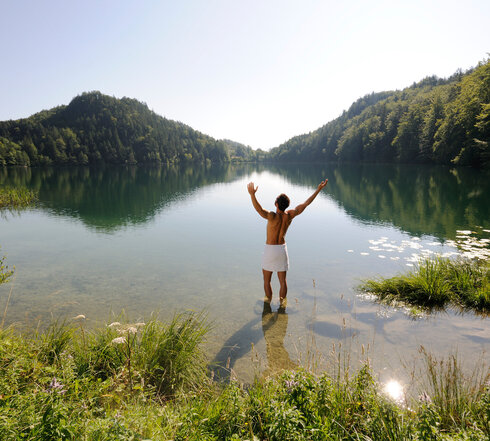 Das Bild zeigt einen Mann, der sich nur ein weißen Handtuch um die Hüften gebunden hat. Er hat dem Betrachter den Rücken zugedreht, steht im knöcheltiefen Wasser eines Sees und streckt die Arme nach oben. Der See ist umringt von grünen Wäldern. Der Himmel ist blau. Die Sonne scheint.