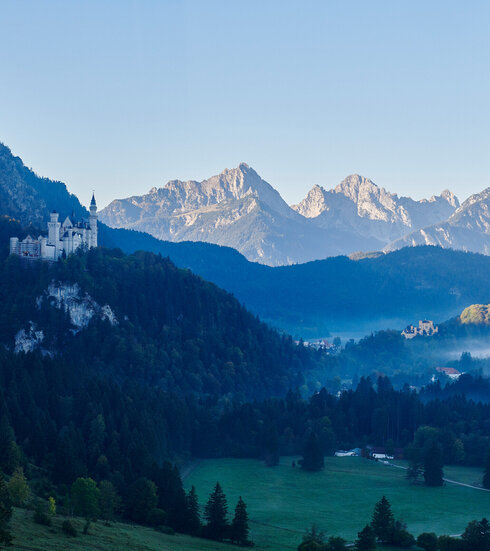 Das Bild zeigt die beiden Schlösser Hohenschwangau und Neuschwanstein, die auf zwei Anhöhen über den umliegenden Wiesen und Wäldern thronen. Im Hintergrund erheben sich die Berge. Der Himmel ist blau. Die Sonne geht gerade auf. Die Schlösser liegen noch im Schatten. 