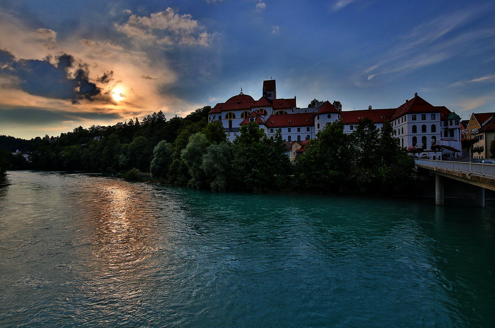Das Bild zeigt ein schlossähnliches Gebäude, das auf einer Anhöhe am Ufer des Flusses Lech thront. Bei dem Gebäudekomplex handelt es sich um das ehemalige Benediktinerkloster St. Mang, das mit seinem roten Dach, majestätisch vor dem Hintergrund eines klaren blauen Himmels steht. Der türkise Fluss, fließt sanft vor dem Gebäude und spiegelt das Sonnenlicht. Die umgebende Landschaft ist üppig grün, mit Bäumen und Büschen, die die Landschaft prägen. Die untergehende Sonne tauch die Szene in warmes Licht.