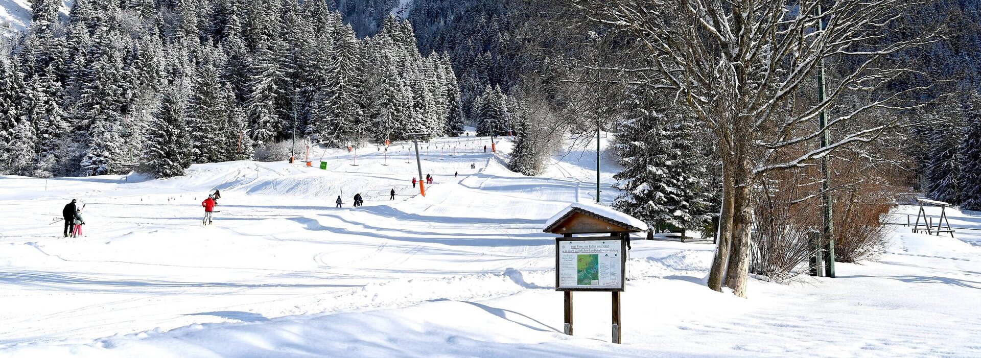 Das Bild zeigt einen hohen Berg. Auf einer Anhöhe davor ist ein Skilift samt Piste zu sehen. Zahlreiche Menschen stehen im Schlepplift und lassen sich nach oben fahren. Alles ist schneebedeckt. Der Himmel ist wolkenverhangen. Die Sonne scheint.  