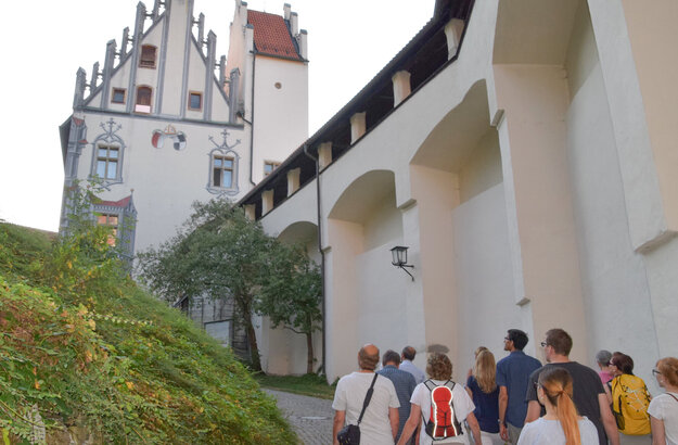 Das Bild zeigt eine Gruppe Menschen, die dem Betrachter weggewandt sind. Die Gruppe steht im Schlosshof des Hohen Schlosses und laufen in Richtung des Hauptgebäudes. Die Abendsonne taucht die Szene in ein warmes Licht. 