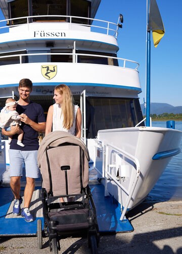 Das Bild fängt eine friedvolle Szene auf einem Schiff ein. Ein Mann und eine Frau stehen an Deck des Schiffs, das an einer Pier festgemacht ist und scheinen, als möchten sie von Borg gehen. Der Mann hält ein Baby in seinen Armen und die Frau steht neben ihm, einen Kinderwagen schiebend. Das Schiff ist weiß und blau und es weht eine Deutschland-Flagge im Wind, die am Schiff befestigt ist. Die Pier erstreckt sich ins türkisblaue Wasser des Forggensees. Im Hintergrund sind Bäume und Berge zu sehen. 