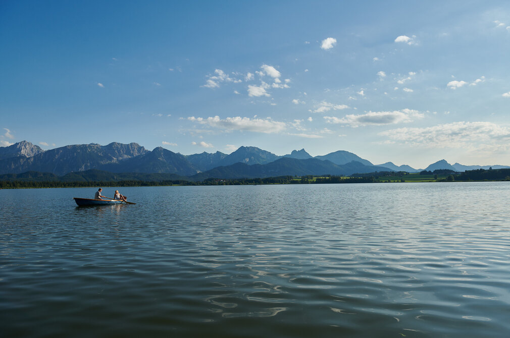 Das Bild zeigt eine zwei Menschen, die in einem Boot über den ruhigen Hopfensee rudern. Der See ist von malerischen Bergen umgeben, deren Gipfel in den Himmel ragen. Das Boot mit seinen beiden Insassen befindet sich in der Mitte des Sees und erzeugt so ein Gefühl von Ausgeglichenheit und Harmonie mit der Natur. Die Ruhe des Wassers wird im Himmel über dem See widergespiegelt, wo wenige Wolken zerstreut sind und die weite Fläche des blauen Himmels mit Tiefe bereichern. Die Gesamtkomposition des Bildes suggeriert einen friedlichen Tag in Begleitung der Natur.