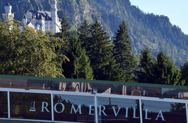 Das Bild zeigt Schloss Neuschwanstein. Im Vordergrund ist ein Teil eines anderen Gebäudes zu sehen auf dem die Aufschrift Römervilla zu lesen ist. 