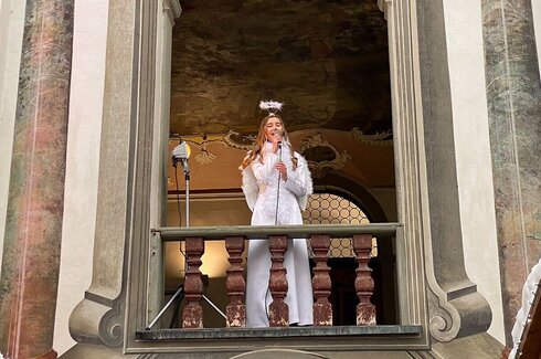 Das Bild ist ein Foto einer Person, die auf einem Balkon steht. Der Balkon ist Teil eines aufwendig gestalteten Gebäudes mit klassischen architektonischen Merkmalen, wie Säulen und einem dekorativen Geländer. Die Person trägt ein weißes Kleid und ein weißes Kopfschmuckstück, das einem Heiligenschein ähnelt. Sie hält ein Mikrofon und scheint zu sprechen oder zu singen. Der Hintergrund des Bildes zeigt einen Innenraum mit einer hohen Decke und einem großen, ornamentierten Fenster. Die Beleuchtung im Foto ist weich und diffus, was auf einen Innenraum mit natürlichem Licht durch das Fenster hindeutet.