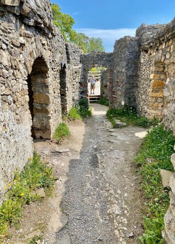  Das Bild zeigt eine alte Steinstruktur, möglicherweise eine Burg oder Festung, mit einer Person im Zentrum eines Innenhofs. Die Struktur ist aus großen, unregelmäßig geformten Steinen gebaut, und die Wände sind mit Moos und Vegetation bedeckt, was auf ihr Alter und die Einwirkung der Elemente hinweist. Der Innenhof wird von einer Steinmauer umgeben, und es sind mehrere Bögen und Tore sichtbar. Die Person im Innenhof trägt ein weißes Hemd und dunkle Hosen und scheint sich in Richtung der Kamera zu wenden. Der Himmel ist klar und blau, was auf einen sonnigen Tag hinweist. Die Gesamtatmosphäre des Bildes ist die Bedeutung historischer Bedeutung und ein Gefühl der Einsamkeit.