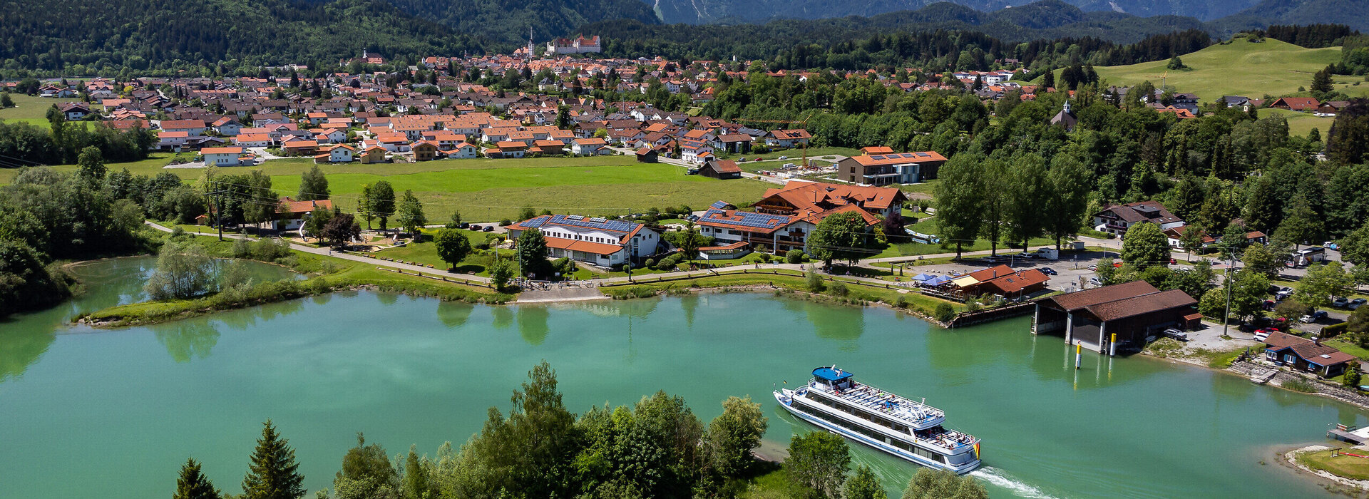 Das Foto zeigt einen Blick auf die Stadt Füssen vom türkisen Forggensee aus. Im Hintergrund zieht sich die Alpenkette durch das Foto. zu sehen. Auf einer Anhöhe vor den Bergen thront das Hohe Schloss über Füssen. Es ist Sommer. Der Himmel ist blau. Die Sonne scheint.