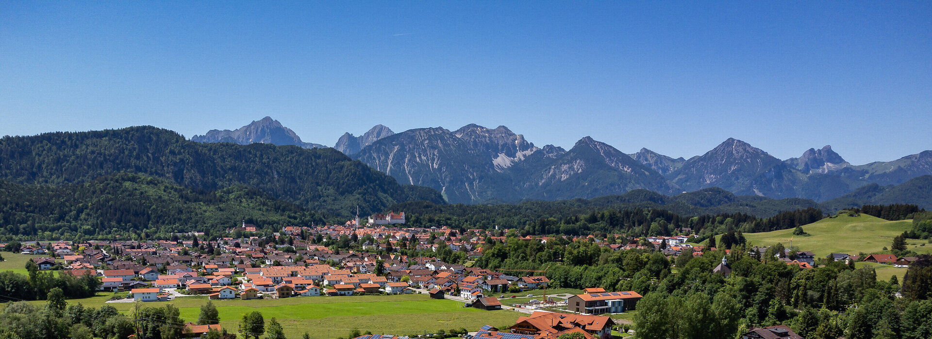Das Foto zeigt einen Blick auf die Stadt Füssen vom türkisen Forggensee aus. Im Hintergrund zieht sich die Alpenkette durch das Foto. zu sehen. Auf einer Anhöhe vor den Bergen thront das Hohe Schloss über Füssen. Es ist Sommer. Der Himmel ist blau. Die Sonne scheint.
