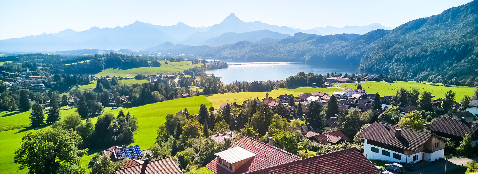 Das Bild den Ort Weißensee von einer Anhöhe aus. Man sieht den gleichnamigen See. Dahinter erheben sich bewaldete Hügel und unzählige Gipfel. Der Himmel ist blau. Die Sonne scheint.  