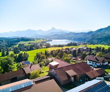 Das Bild den Ort Weißensee von einer Anhöhe aus. Man sieht den gleichnamigen See. Dahinter erheben sich bewaldete Hügel und unzählige Gipfel. Der Himmel ist blau. Die Sonne scheint.  