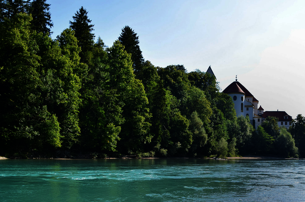 Das Bild zeigt ein Paar, das an einem steinigen Ufer des Flusses Lech steht. Der Mann und die Frau stehen dicht beieinander, ihre Körper sind dem Wasser zugewandt. Der türkise Fluss liegt im Fokus des Bildes. Auf der gegenüberliegenden Seite erhebt sich ein großer Gebäudekomplex mit weißen Mauern und einem roten Dach. Hierbei handelt es sich um das Hohe Schloss und das ehemalige Kloster St. Mang in Füssen. Der Himmel darüber ist klar, blau und wolkenlos.