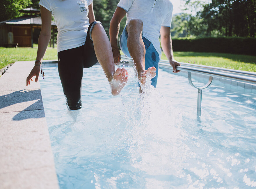 Das Bild zeigt zwei Personen, die in einem Kneipptretbecken stehen. Man sieht nicht ihre Gesichter. Beide Personen heben ein Bein aus dem Wasser, sodass die Wassertropfen um sie herum spritzen. im Hintergrund zeigt sich ein grüner Park mit satten Wiesen und belaubten Bäumen.