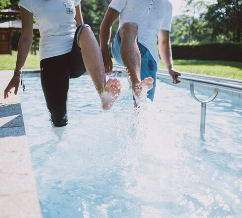 Das Bild zeigt zwei Personen, die in einem Kneipptretbecken stehen. Man sieht nicht ihre Gesichter. Beide Personen heben ein Bein aus dem Wasser, sodass die Wassertropfen um sie herum spritzen. im Hintergrund zeigt sich ein grüner Park mit satten Wiesen und belaubten Bäumen.