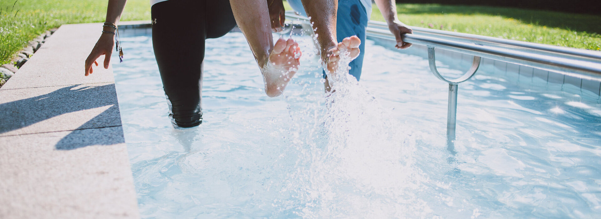 Das Bild zeigt zwei Personen, die in einem Kneipptretbecken stehen. Man sieht nicht ihre Gesichter. Beide Personen heben ein Bein aus dem Wasser, sodass die Wassertropfen um sie herum spritzen. im Hintergrund zeigt sich ein grüner Park mit satten Wiesen und belaubten Bäumen.