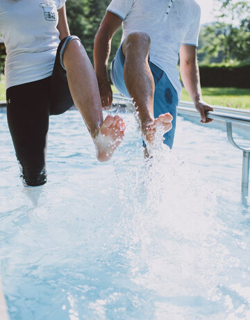 Das Bild zeigt zwei Personen, die in einem Kneipptretbecken stehen. Man sieht nicht ihre Gesichter. Beide Personen heben ein Bein aus dem Wasser, sodass die Wassertropfen um sie herum spritzen. im Hintergrund zeigt sich ein grüner Park mit satten Wiesen und belaubten Bäumen.