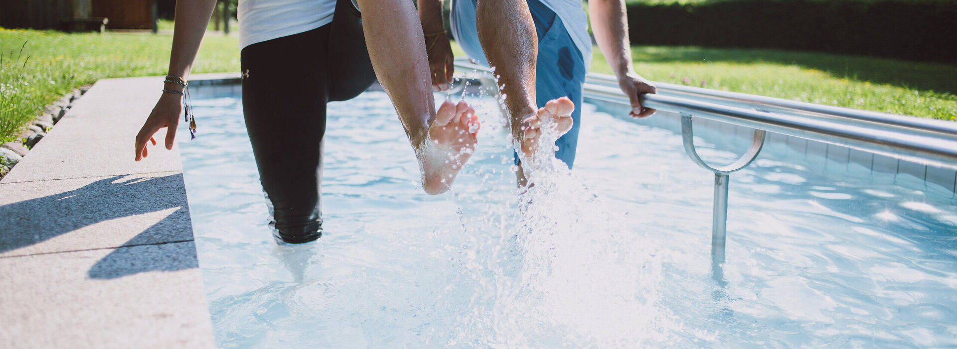Das Bild zeigt zwei Personen, die in einem Kneipptretbecken stehen. Man sieht nicht ihre Gesichter. Beide Personen heben ein Bein aus dem Wasser, sodass die Wassertropfen um sie herum spritzen. im Hintergrund zeigt sich ein grüner Park mit satten Wiesen und belaubten Bäumen.
