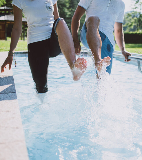 Das Bild zeigt zwei Personen, die in einem Kneipptretbecken stehen. Man sieht nicht ihre Gesichter. Beide Personen heben ein Bein aus dem Wasser, sodass die Wassertropfen um sie herum spritzen. im Hintergrund zeigt sich ein grüner Park mit satten Wiesen und belaubten Bäumen.