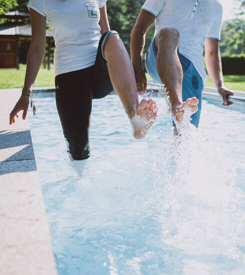 Das Bild zeigt zwei Personen, die in einem Kneipptretbecken stehen. Man sieht nicht ihre Gesichter. Beide Personen heben ein Bein aus dem Wasser, sodass die Wassertropfen um sie herum spritzen. im Hintergrund zeigt sich ein grüner Park mit satten Wiesen und belaubten Bäumen.