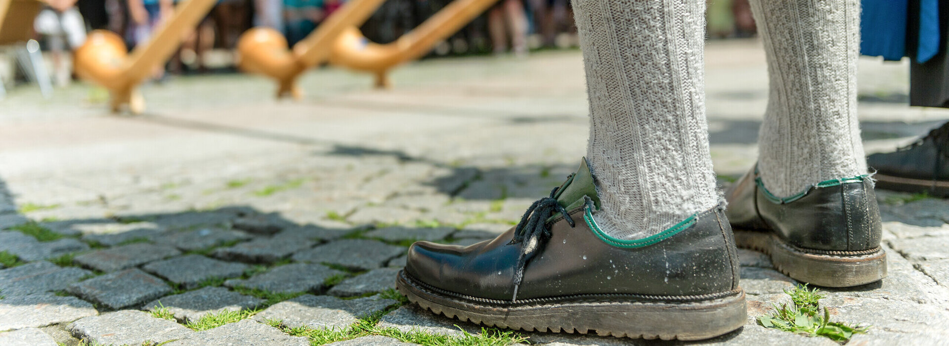 Das Bild zeigt mehrere Alphornspieler. Allerdings sind nur zwei Beine bis zu den Knien zu sehen. Die Beine sind mit grauen Kniestrümpfen und schwarzen Haferlschuhen bekleidet. Um die Alphörner kann man mehrere Zuschauer erkennen. Die Sonne scheint. Die Szenerie findet wohl auf einem Platz in der Stadt statt. Im Hintergrund sind Hausfassaden zu erkennen. Auf dem Boden liegt Kopfsteinpflaster.   