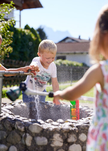 Das Bild zeigt eine lebhafte Szene von drei Kindern, die im Garten spielen. Die Kinder, zwei Jungen und ein Mädchen, sind in ihr Spiel vertieft. Das Mädchen, das ein pinkes Kleid trägt, steht neben einem Wasserfass, seine Hand greift aus, um es anzustellen. Das Wasserfass, das zentrale Feature im Bild, wird von Steinen umgeben, was der Szene einen Hauch der Natur verleiht.  Die beiden Jungen, einer in einem gelben Hemd und der andere in einem weißen Hemd, stehen auf der anderen Seite des Fasses. Sie halten einen Schlauch, bereit, Wasser zu sprühen und sich an der Spielfreude zu beteiligen. Der Garten ist ein üppiges Grün, mit Bäumen und Büschen, die das Bild beleben.  Im Hintergrund ist ein Haus zu sehen, was darauf hindeutet, dass diese spielerische Szene in einem Wohngebiet stattfindet. Das Bild ist ein Schnappschuss von Kinderfreude und Unschuld, der einen Moment ungezügelten Vergnügens einfängt.